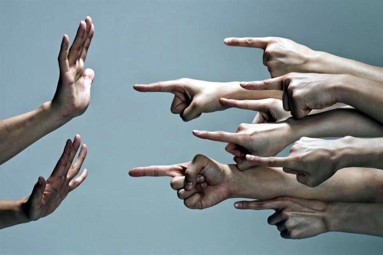 Imagen simbólica donde muchas manos señalan con el dedo indice a una persona que esta sobre la izquierda, solo se ven las manos levantadas en modo defensivo.