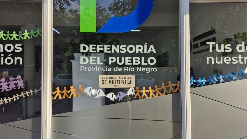 Ventana del edificio de la defensoria con un cartel hecho en papel que contiene la frase "la memoria en marcha se multiplica" y figuras de personas unidas de la mano.