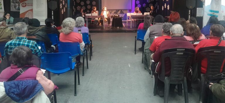 Publico de personas sentados en sillas escuchando a los oradores al frente sobre un escenario.